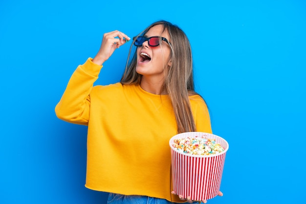 Jeune femme de race blanche isolée sur fond bleu avec des lunettes 3D et tenant un grand seau de pop-corn