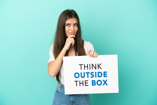 Jeune femme de race blanche isolée sur fond bleu tenant une pancarte avec du texte Think Outside The Box et la pensée