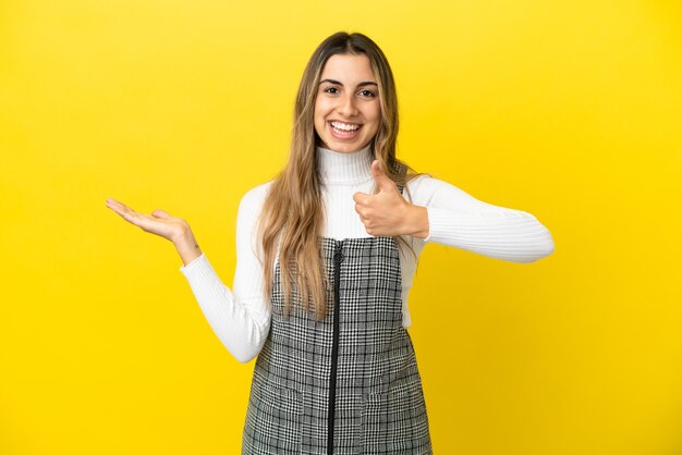 Jeune femme de race blanche isolée sur fond jaune tenant copyspace imaginaire sur la paume pour insérer une annonce et avec les pouces vers le haut