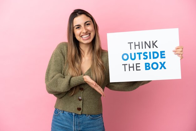 Jeune femme de race blanche isolée sur fond rose tenant une pancarte avec du texte Think Outside The Box et le pointant