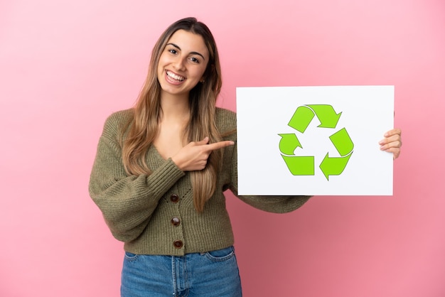 Jeune femme de race blanche isolée sur fond rose tenant une pancarte avec l'icône de recyclage et la pointant