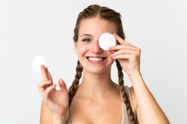 Jeune femme de race blanche isolée sur un mur blanc avec un coton pour enlever le maquillage de son visage et souriant