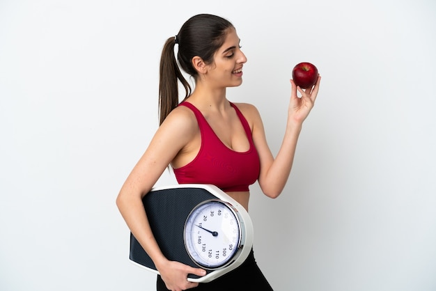 Jeune femme de race blanche isolée sur un mur blanc avec une machine à peser et avec une pomme