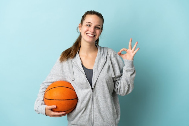 Jeune femme de race blanche isolée sur mur bleu jouant au basket et faisant signe OK