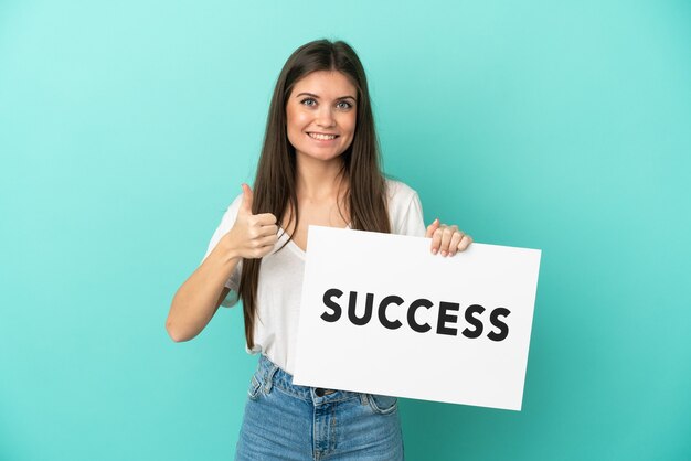 Jeune femme de race blanche isolée sur un mur bleu tenant une pancarte avec texte SUCCESS avec le pouce vers le haut