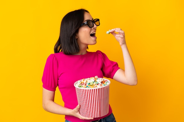 Jeune femme de race blanche isolée sur un mur jaune avec des lunettes 3D et tenant un grand seau de pop-corn