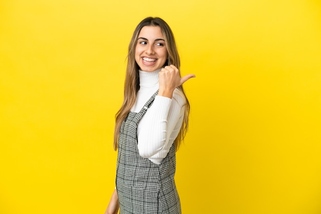 Jeune femme de race blanche isolée sur un mur jaune pointant vers le côté pour présenter un produit