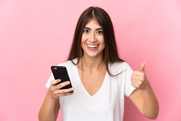 Jeune femme de race blanche isolée sur un mur rose à l'aide de téléphone mobile tout en faisant les pouces vers le haut