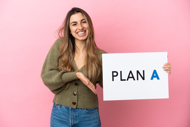 Jeune femme de race blanche isolée sur un mur rose tenant une pancarte avec le message PLAN A et le pointant