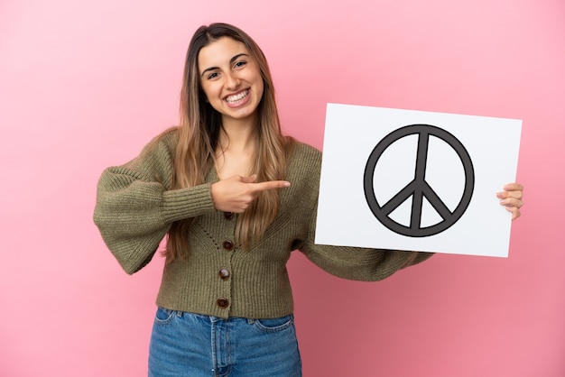 Jeune femme de race blanche isolée sur un mur rose tenant une pancarte avec le symbole de la paix et en le pointant