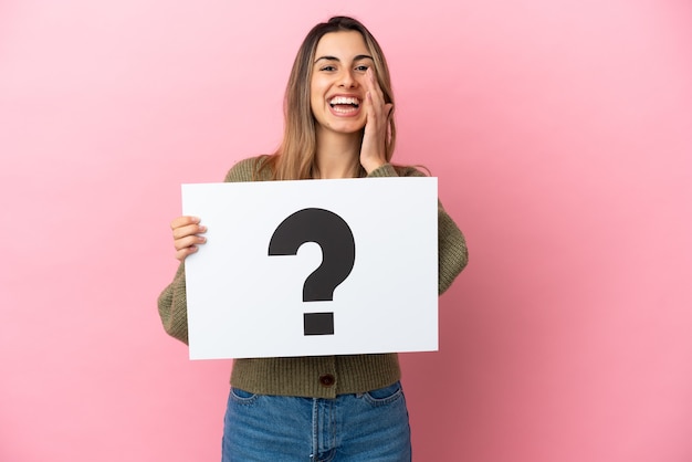 Jeune femme de race blanche isolée sur un mur rose tenant une pancarte avec le symbole de point d'interrogation et criant