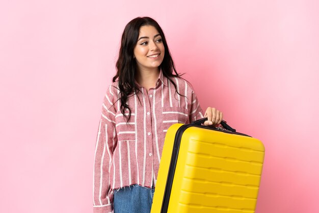 Jeune femme de race blanche isolée sur un mur rose en vacances avec valise de voyage