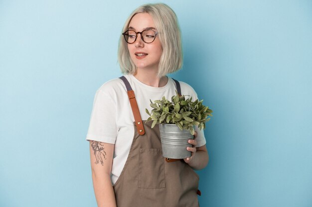 Jeune femme de race blanche jardinier tenant une plante isolée sur fond bleu regarde de côté souriant, joyeux et agréable.