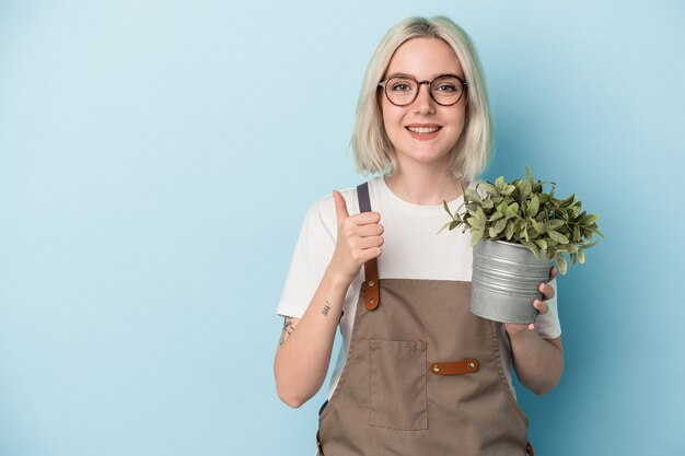 Jeune femme de race blanche jardinier tenant une plante isolée sur fond bleu souriant et levant le pouce vers le haut