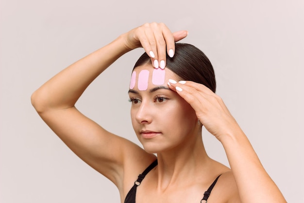 Une jeune femme de race blanche met du ruban kinésio rose sur son visage pour réduire les rides sur son front
