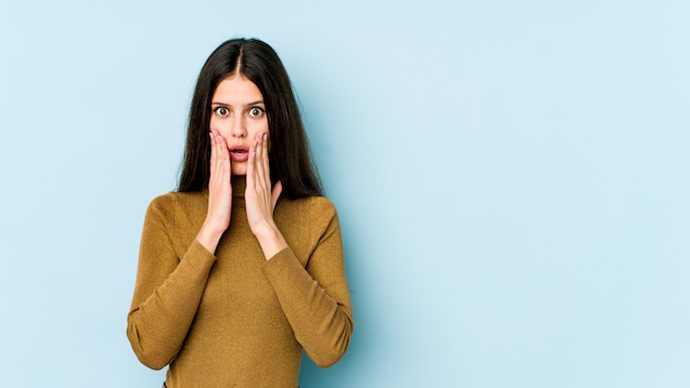 Jeune femme de race blanche sur le mur bleu choqué, couvrant la bouche avec les mains, désireux de découvrir quelque chose de nouveau.