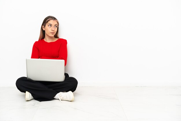 Jeune femme de race blanche avec un ordinateur portable assis sur le sol faisant le geste de doutes