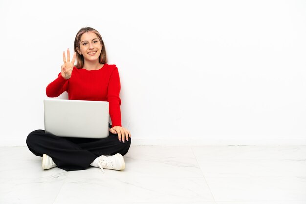 Jeune femme de race blanche avec un ordinateur portable assis sur le sol heureux et comptant trois avec les doigts