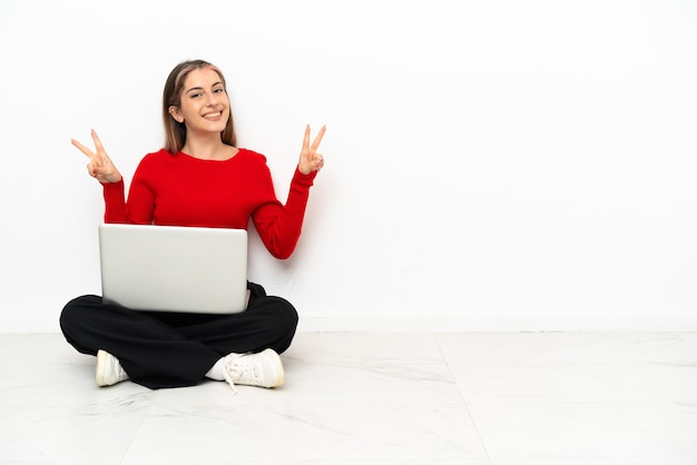 Jeune femme de race blanche avec un ordinateur portable assis sur le sol montrant le signe de la victoire avec les deux mains