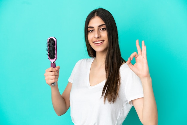 Jeune femme de race blanche avec un peigne à cheveux isolé sur fond bleu montrant un signe ok avec les doigts