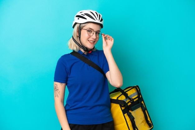 Jeune femme de race blanche avec sac à dos thermique isolé sur fond bleu avec des lunettes et heureux