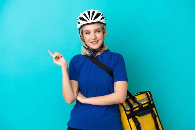 Jeune femme de race blanche avec sac à dos thermique isolé sur fond bleu, pointant le doigt sur le côté