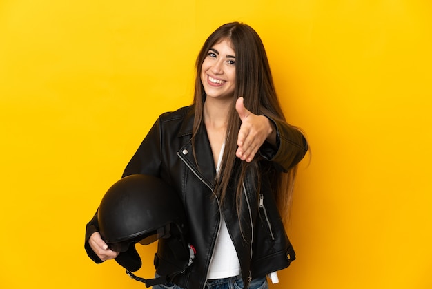Jeune femme de race blanche tenant un casque de moto isolé sur fond jaune se serrant la main pour conclure une bonne affaire