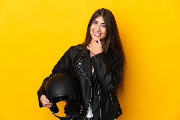 Jeune femme de race blanche tenant un casque de moto isolé sur un mur jaune à la recherche sur le côté et souriant