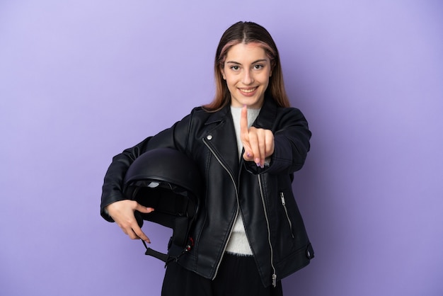 Jeune femme de race blanche tenant un casque de moto isolé sur un mur violet montrant et en soulevant un doigt