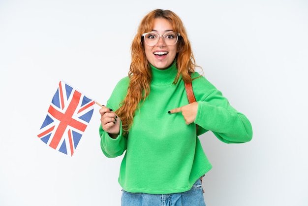 Jeune femme de race blanche tenant un drapeau du Royaume-Uni isolé sur fond blanc avec une expression faciale surprise