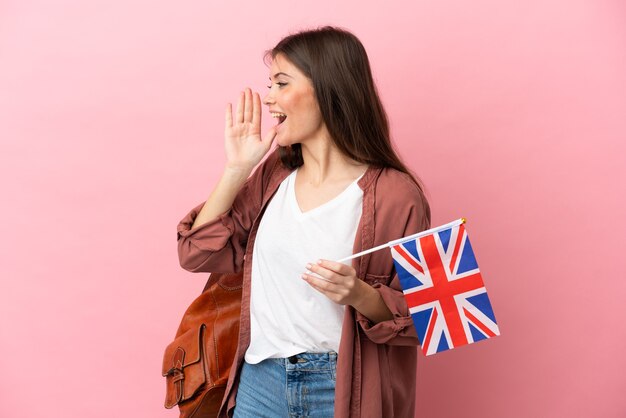 Jeune femme de race blanche tenant un drapeau du Royaume-Uni isolé sur fond rose en criant avec la bouche grande ouverte sur le côté