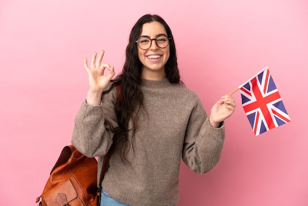 Jeune femme de race blanche tenant un drapeau du Royaume-Uni isolé sur fond rose montrant un signe ok avec les doigts