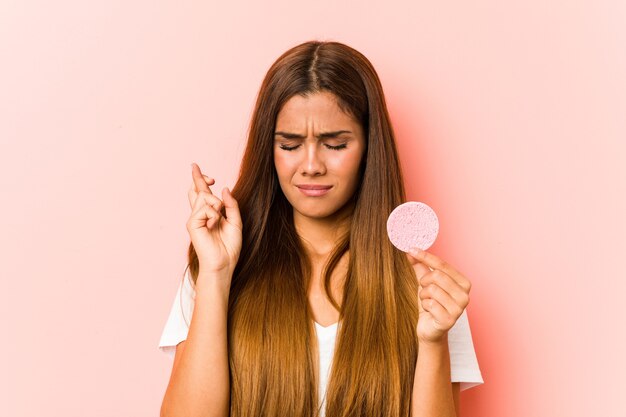 Jeune femme de race blanche tenant une éponge faciale croisant les doigts pour avoir de la chance