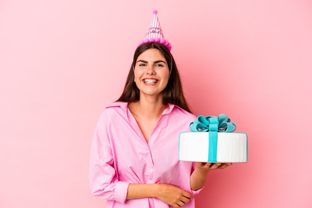 Jeune femme de race blanche tenant un gâteau pour célébrer un anniversaire isolé sur un mur rose en riant et en s'amusant