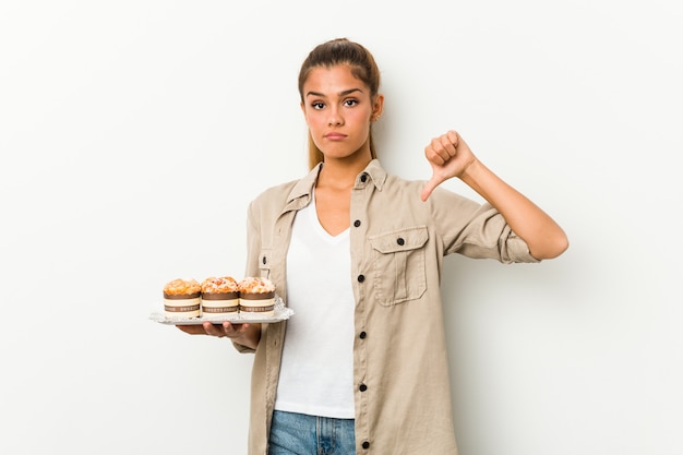 Jeune Femme De Race Blanche Tenant Des Gâteaux Sucrés Montrant Un Geste D'aversion, Les Pouces Vers Le Bas. Concept De Désaccord.