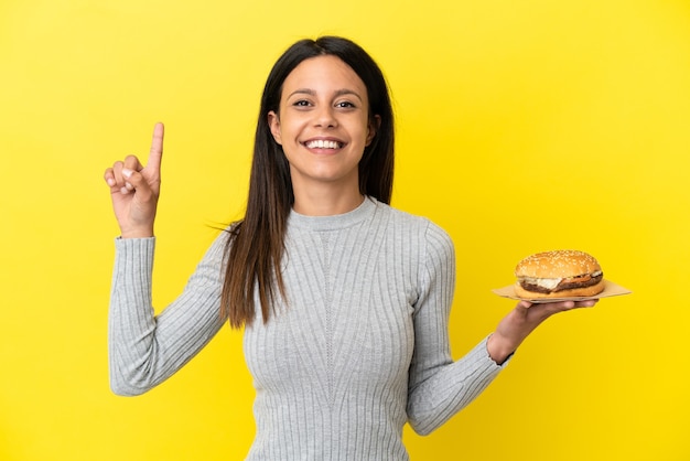 Jeune femme de race blanche tenant un hamburger isolé sur fond jaune pointant vers une excellente idée