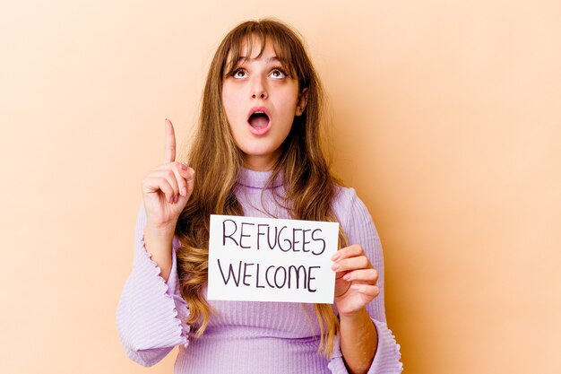 Jeune femme de race blanche tenant une pancarte de bienvenue de réfugiés isolé pointant vers le haut avec la bouche ouverte.