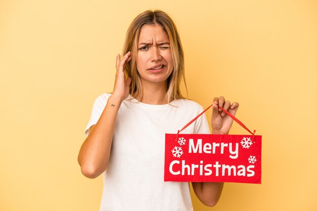 Jeune femme de race blanche tenant une pancarte joyeux noël isolée sur fond jaune couvrant les oreilles avec les mains.