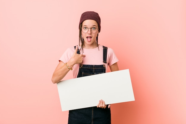 Jeune femme de race blanche tenant une pancarte surprise pointant le doigt sur lui-même, souriant largement.