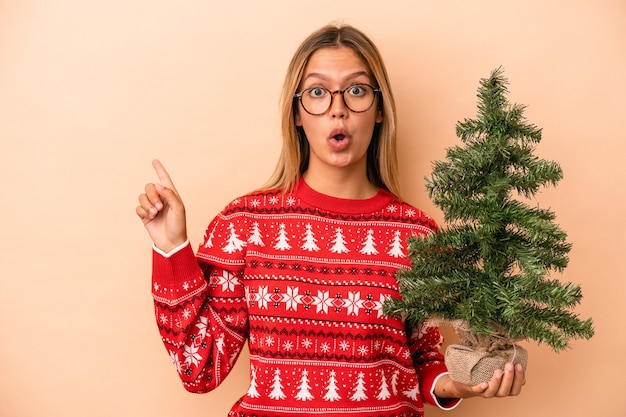 Jeune femme de race blanche tenant un petit arbre de Noël isolé sur fond beige pointant vers le côté