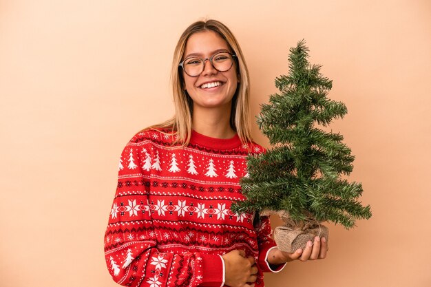 Jeune femme de race blanche tenant un petit arbre de Noël isolé sur fond beige en riant et en s'amusant.