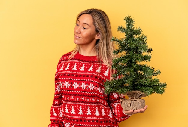 Jeune femme de race blanche tenant un petit arbre de noël isolé sur fond jaune regarde de côté souriant, joyeux et agréable.