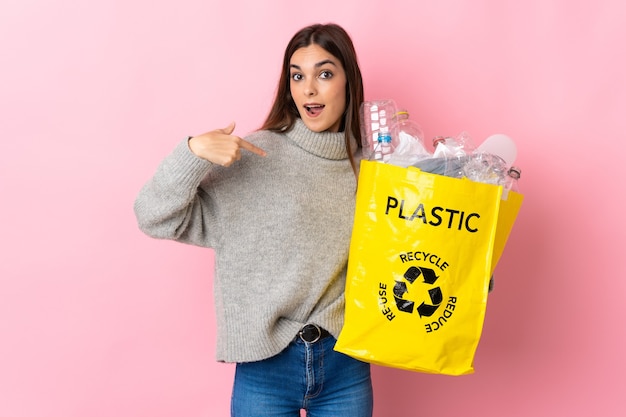 Jeune femme de race blanche tenant un sac plein de bouteilles en plastique à recycler isolé sur rose avec expression faciale surprise