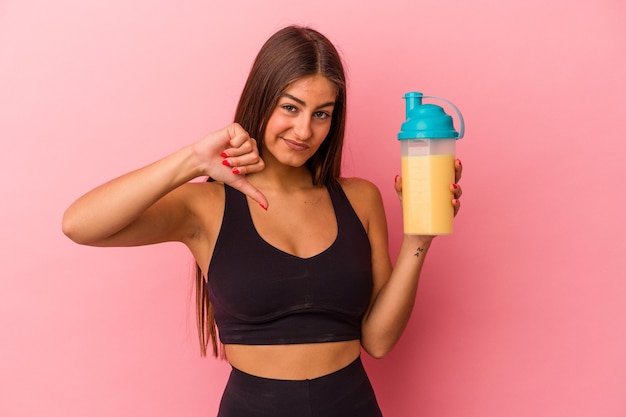 Jeune femme de race blanche tenant un shake protéiné isolé sur fond jaune montrant un geste d'aversion, les pouces vers le bas. Notion de désaccord.