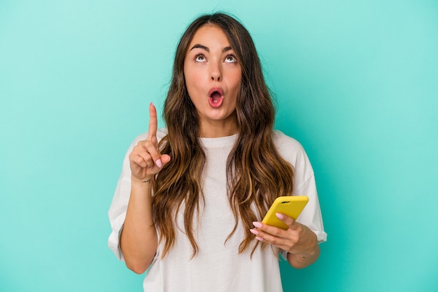 Jeune femme de race blanche tenant un téléphone portable isolé sur fond bleu pointant vers le haut avec la bouche ouverte.