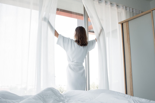 Une jeune femme de race blanche tient des rideaux blancs ouverts à la fenêtre, le matin après s'être réveillée dans la chambre de sa maison. La femme se réveille avec une fraîcheur et ouvre les rideaux aux fenêtres.