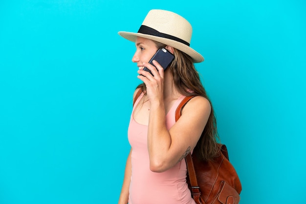 Jeune femme de race blanche en vacances d'été isolée sur fond bleu en gardant une conversation avec le téléphone portable avec quelqu'un