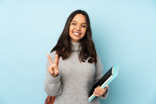 Jeune femme de race mixte allant à l'école isolé sur mur bleu souriant et montrant le signe de la victoire