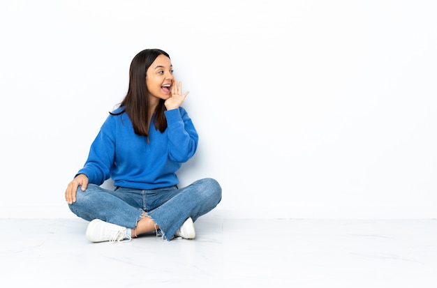 Jeune femme de race mixte assis sur le sol isolé sur blanc criant avec la bouche grande ouverte sur le côté