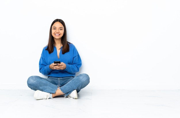 Jeune femme de race mixte assis sur le sol isolé sur blanc en envoyant un message avec le mobile